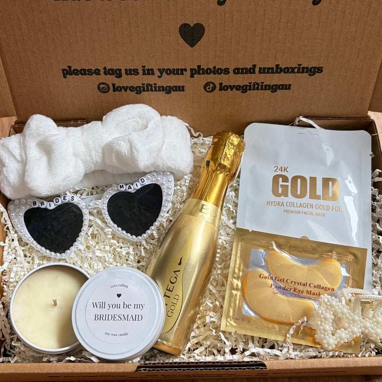 Will you be bridesmaid Gift box with candles, sunglasses, and a bottle of champagne on a wooden surface.
