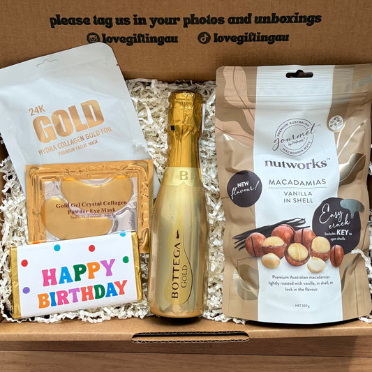 Happy birthday Gift box with a bottle of champagne, snacks, and a card on a wooden surface.