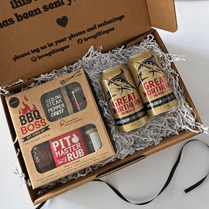 A curated gift box set from Love Gitting featuring a 'BBQ Boss' seasoning set and a 'Pit Master Rub' spice rub in a brown cardboard box with shredded paper, alongside two cans of Great Northern beer.