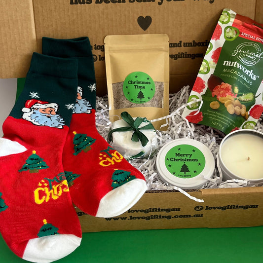 Gifting box with Christmas-themed items including socks, candles, and a bag on a green surface.