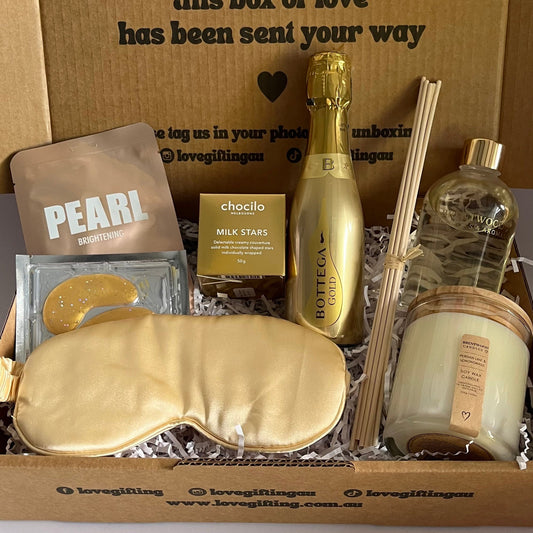 Gift box with items including a bottle of champagne, candles, and a sleep mask on a white background.