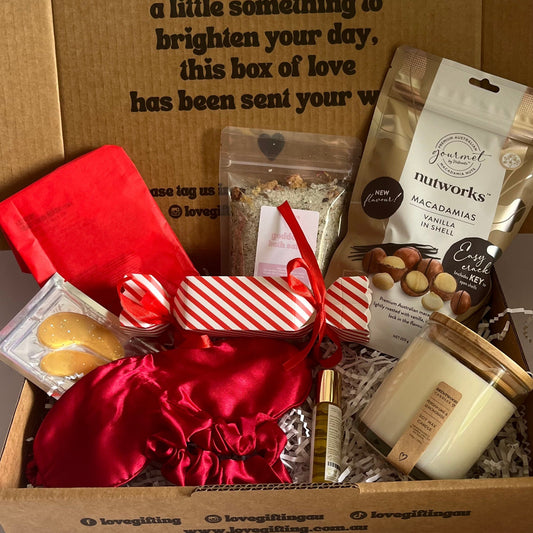 Gift box with various items including a red satin bag, cookies, and a candle on a white surface.