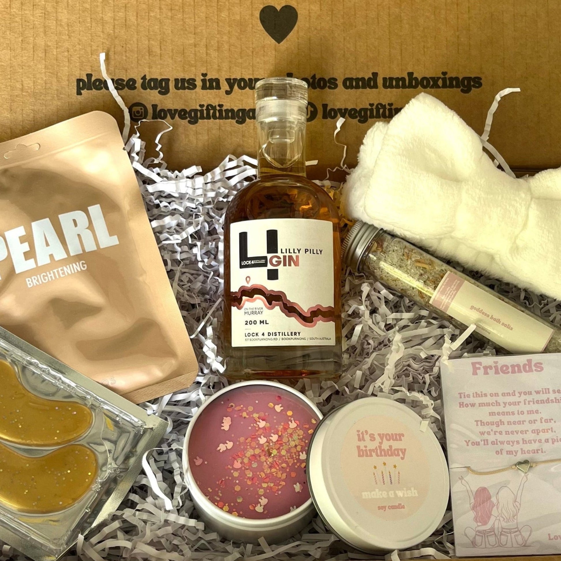 Gift box with various items including a bottle of gin, skincare products, and a candle, with a message on the lid.