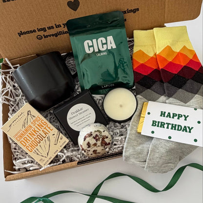 A curated gift box set opened to reveal multiple items: a black candle, a green bag labeled CICA, a small candle tin, a card, a wrapped treat, patterned socks, and a printed message card inside a brown box. A green ribbon is placed in front on a white surface.
