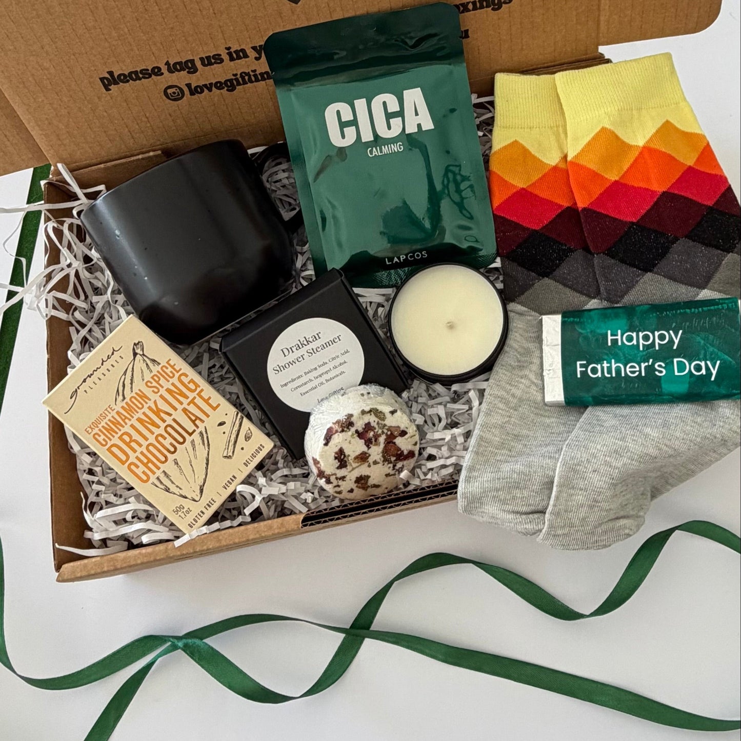 A curated Father's Day gift box filled with various items including a black candle, a green Cica coffee pouch, a book, a chocolate-covered cookie, a pair of multicolor socks, and a gray t-shirt, all arranged in white paper shred inside a brown cardboard box.