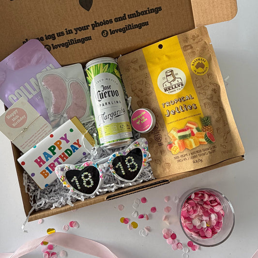 A curated gift box filled with assorted treats and accessories,  the contents include a purple envelope, a can of margarita , margarita lipbalm, Tropical gummies,  18th  sunglasses, 18th bracelet, lapcos facemask , under eye mask and happy birthday chocolate,