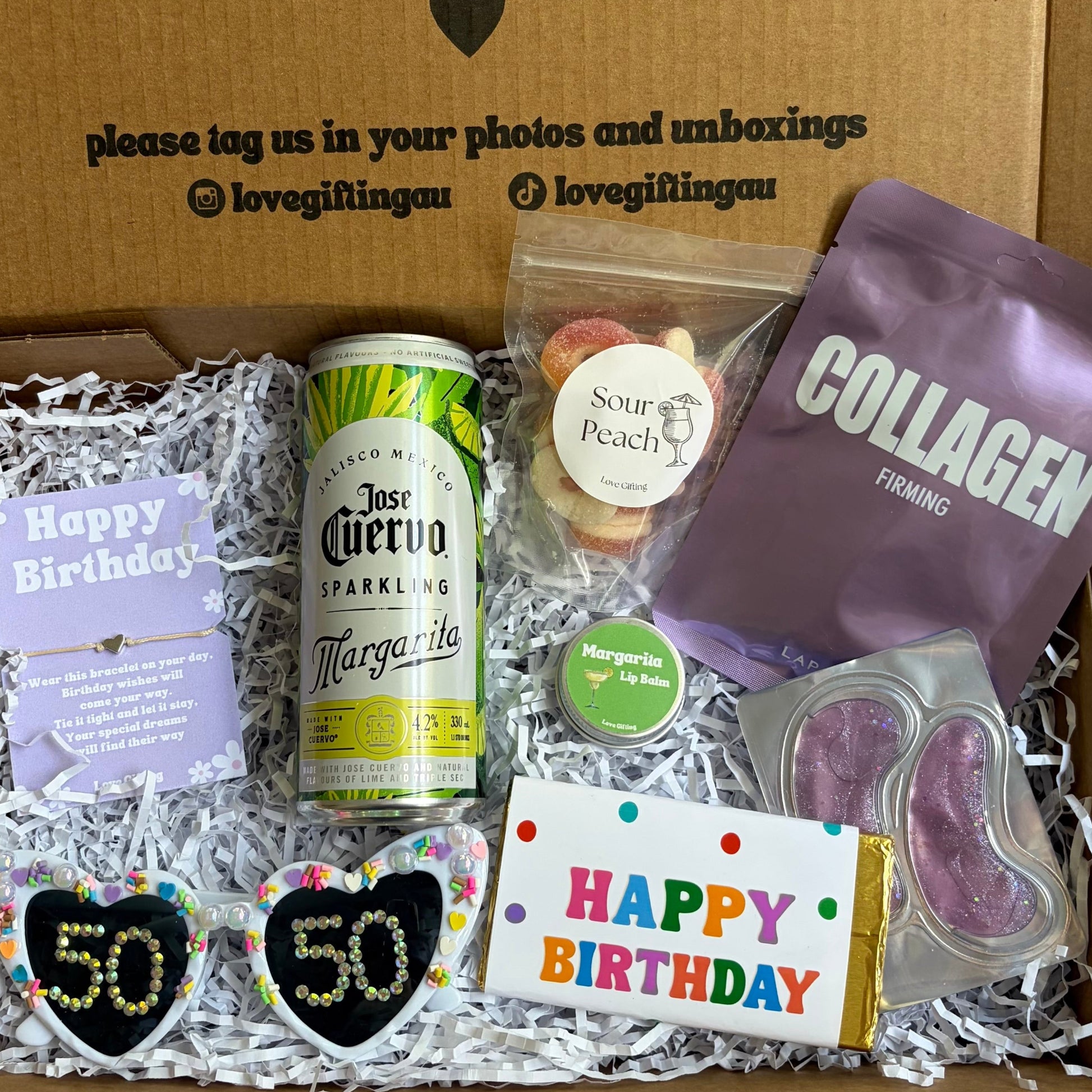 Gift box with birthday-themed items including a bottle, snacks, and a card, with text on the box.