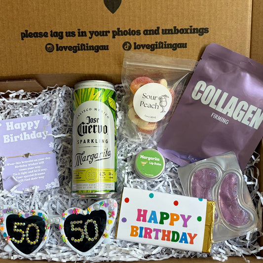 Gift box with birthday-themed items including a bottle, snacks, and a card, with text on the box.