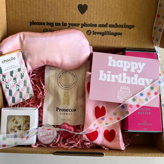 Gift box with birthday-themed items including Prosecco gummies , pink socks, shower steamer, chocolate and cookies 