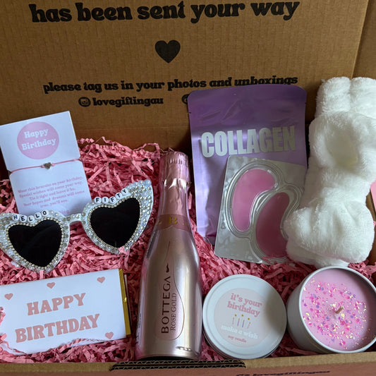 Gift box with birthday-themed items including a bottle, sunglasses, and a card on a pink surface.