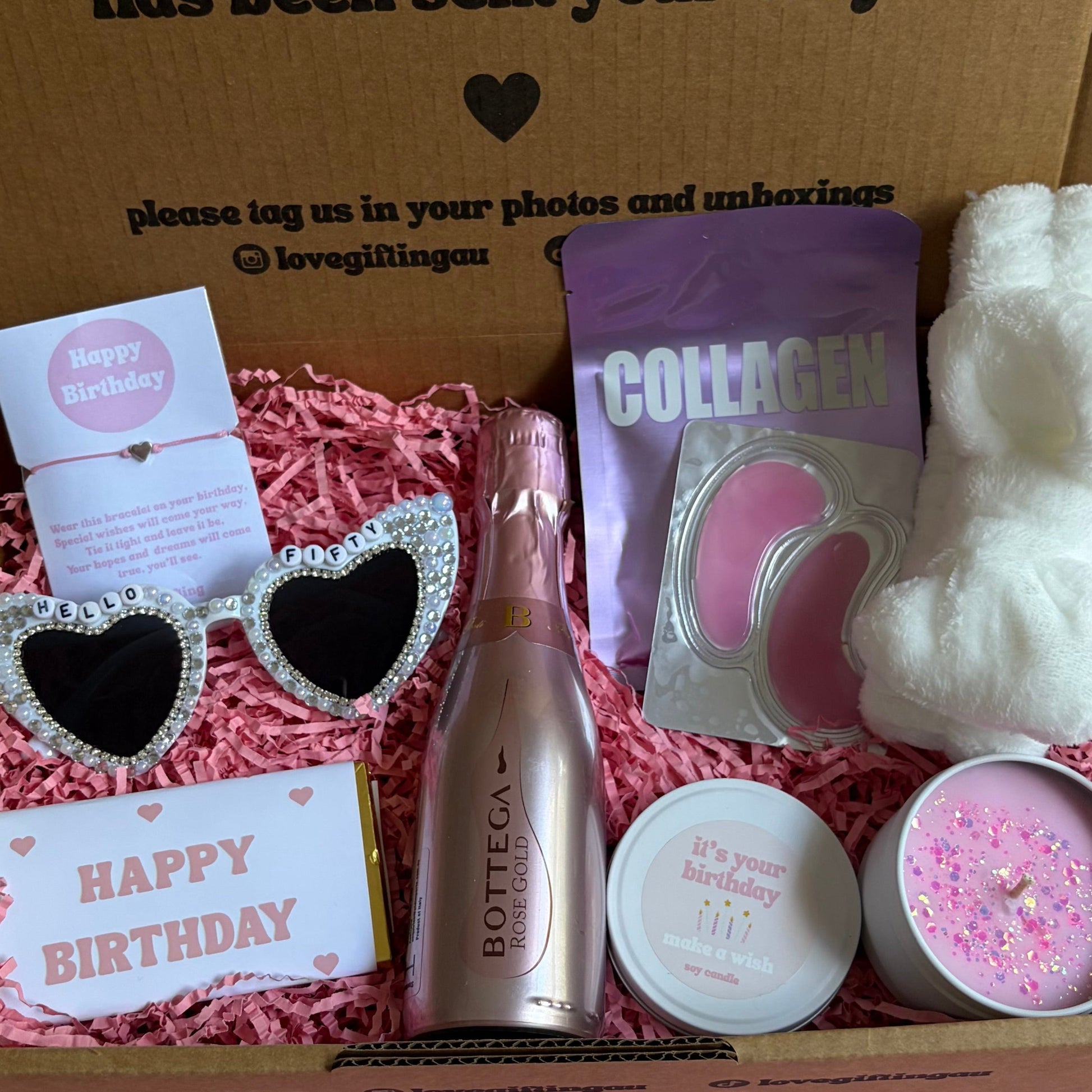 Gift box with birthday-themed items including a bottle, sunglasses, and a card on a pink surface.