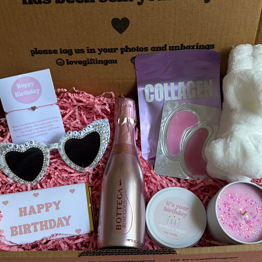 Gift box with birthday-themed items including a bottle, sunglasses, and a card on a pink surface.