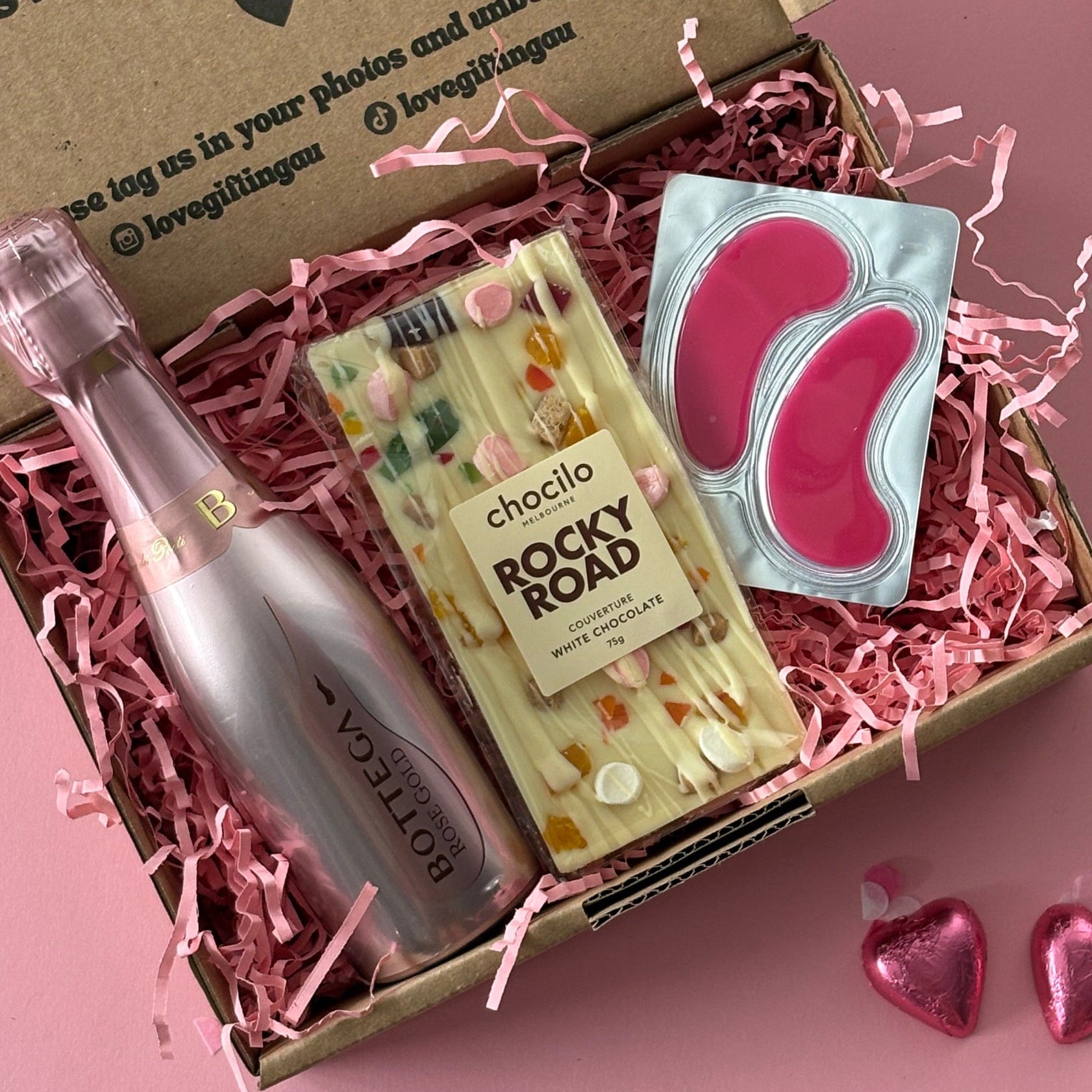 Gift box with a bottle, snacks, and a card on a pink background