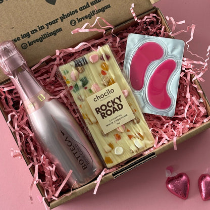 Gift box with a bottle, snacks, and a card on a pink background