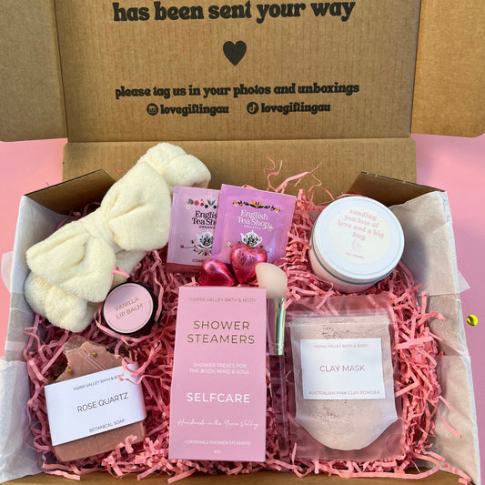 Gift box with self-care items including a candle, soap, and bath bombs on a pink background.