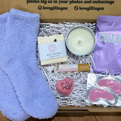 Box of self-care items including socks, a candle, and eye masks on a wooden surface.