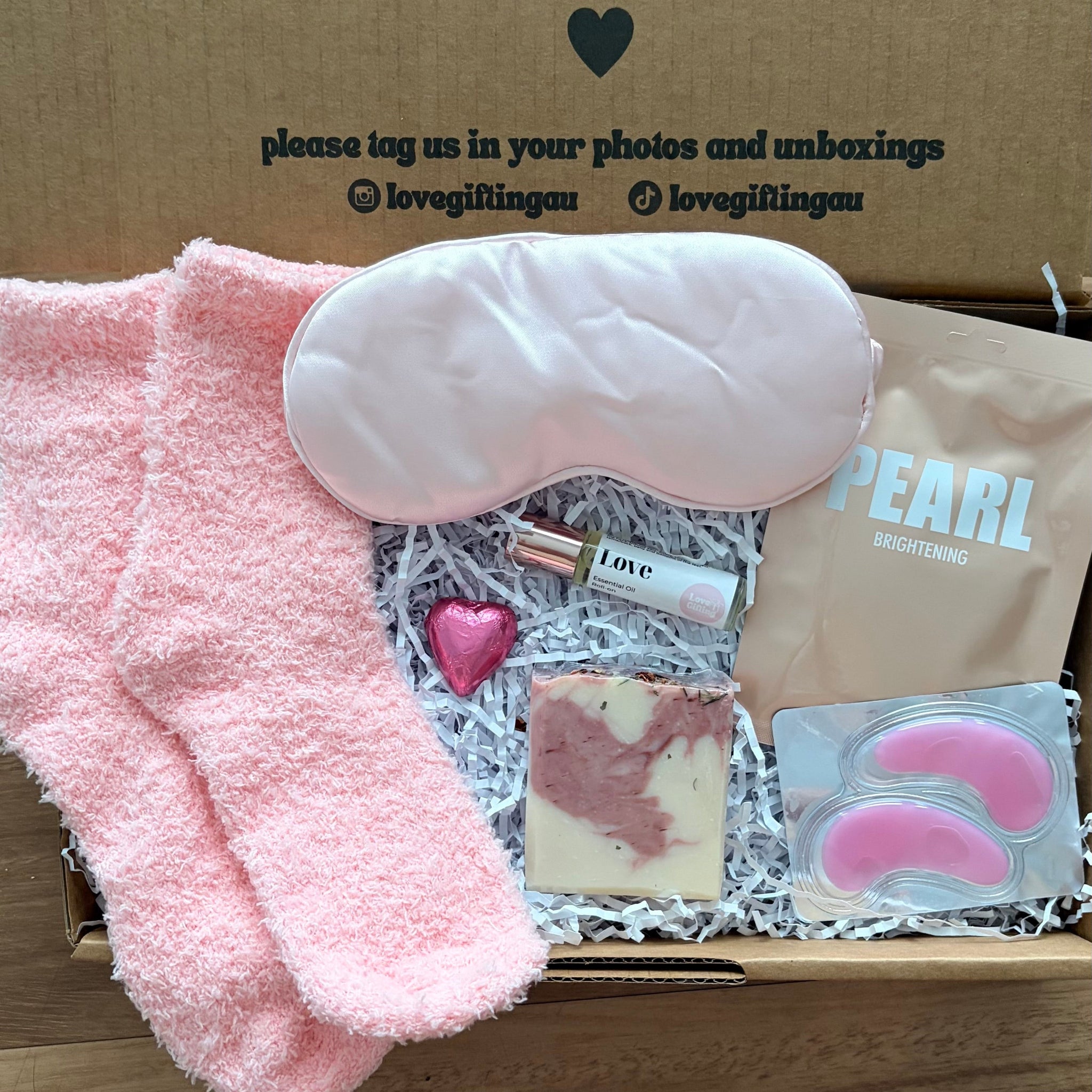 Box of self-care items with a pink eye mask, heart-shaped soap, and pink fuzzy socks on a wooden surface.
