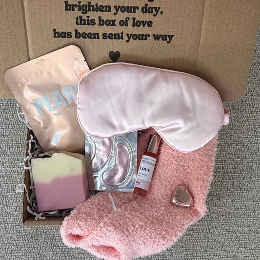 A curated self-care gift box laid open on a gray textured fabric surface, containing a pink plush eye mask, a pink robe, a bar of soap, a sachet, a small bottle, and a printed cardboard insert with a message.