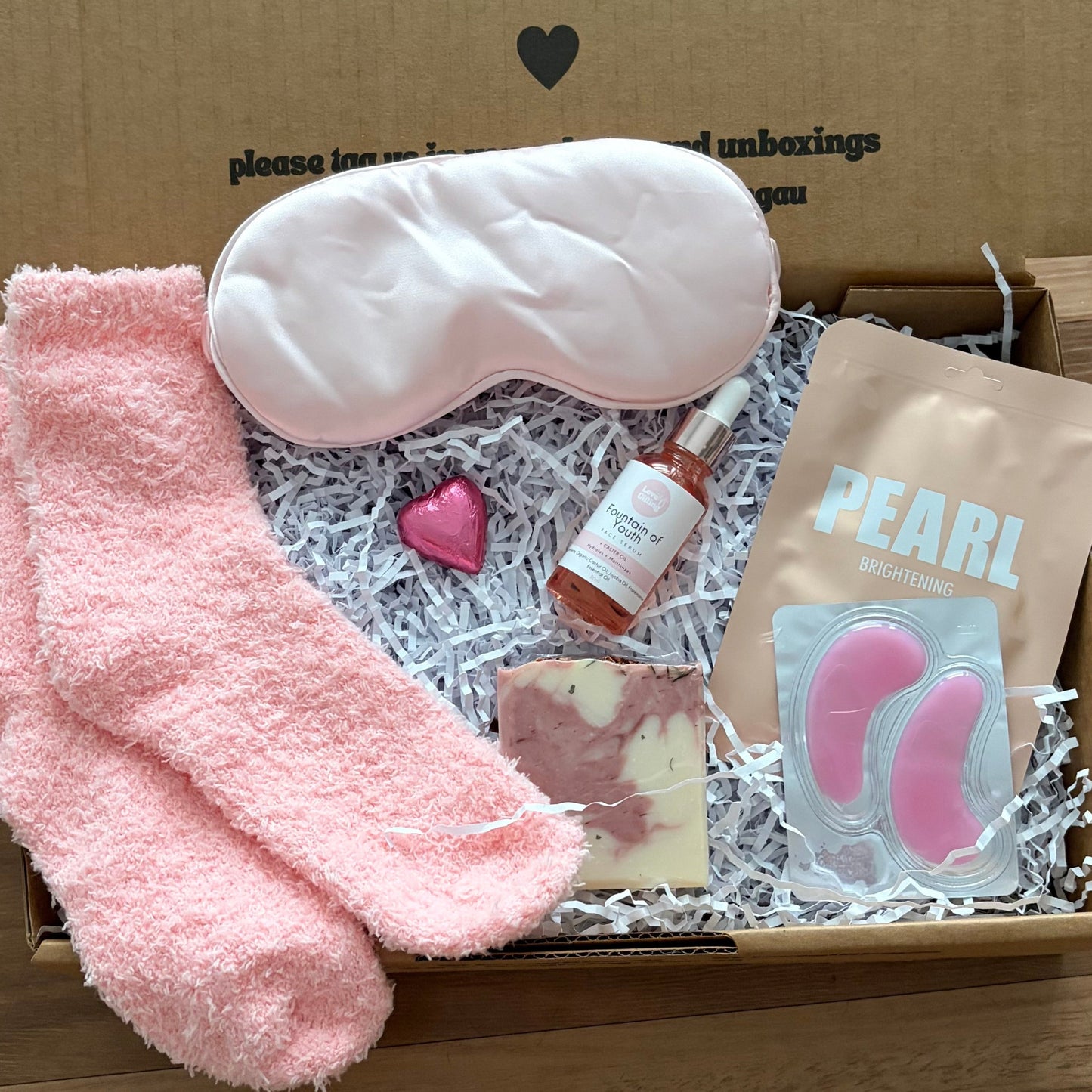 Gift box with pink socks, eye mask, and other items on a wooden surface.