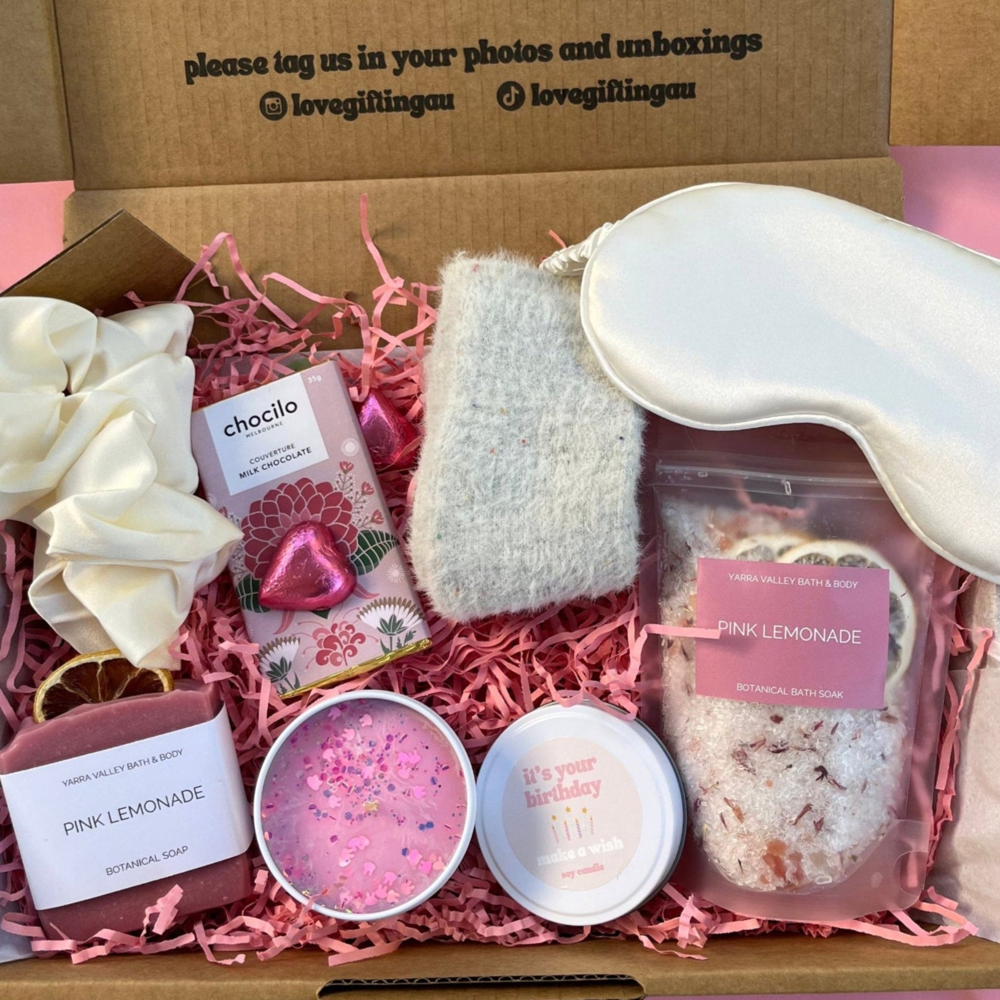 Gift box with self-care items including a silk sleep eye mask, bath soak , satin scrunchie, homemade soap, happy birthday candle, fluffy socks and chocolate.