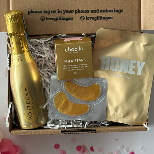 Gourmet gift box with gold items including a bottle, chocolate, and honey on a white background.