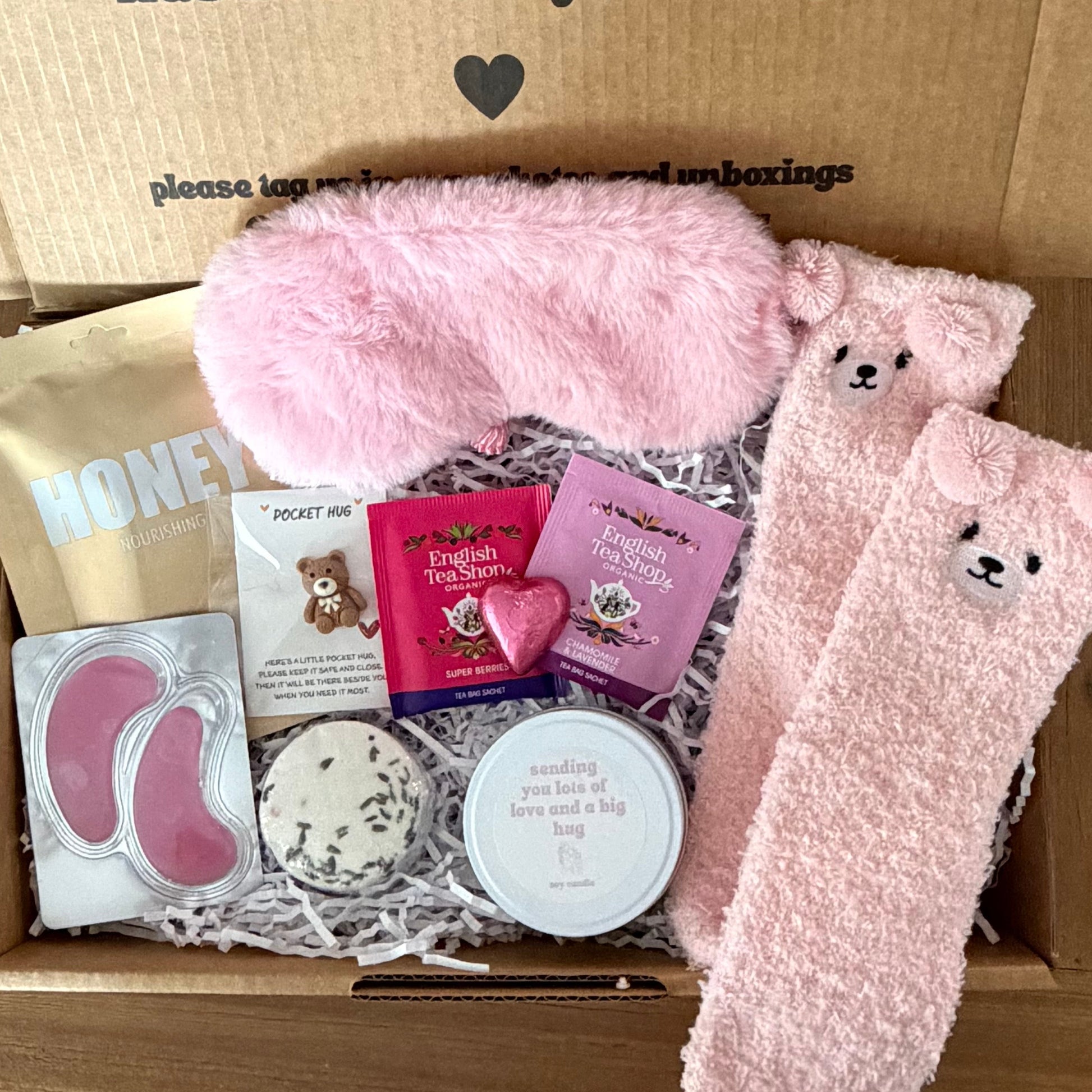 Box of pink items including socks, eye masks, and a heart-shaped box with a message on a wooden floor.