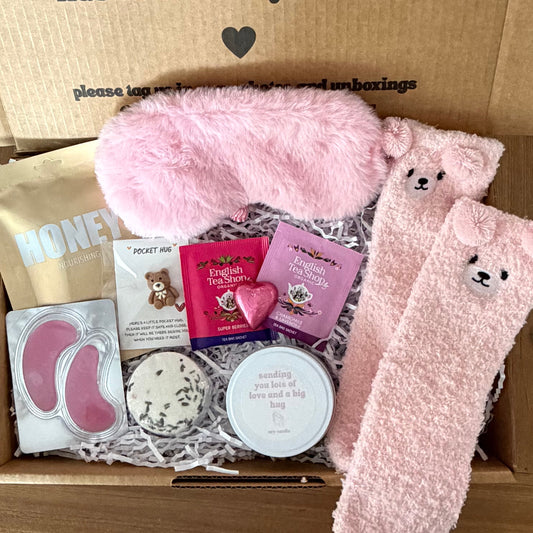 Box of pink items including socks, eye masks, and a heart-shaped box with a message on a wooden floor.