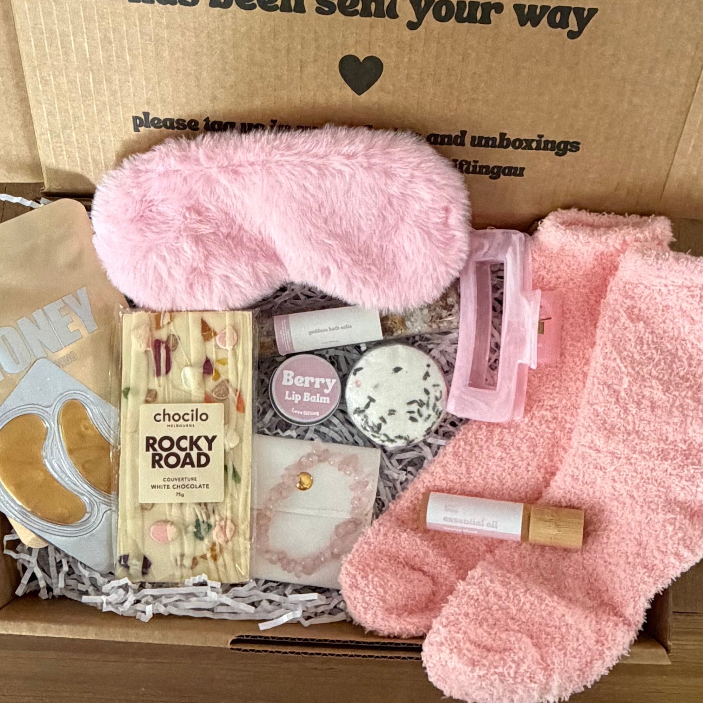 Box of self-care items including pink socks, eye mask, and bath bombs with a message on the lid.