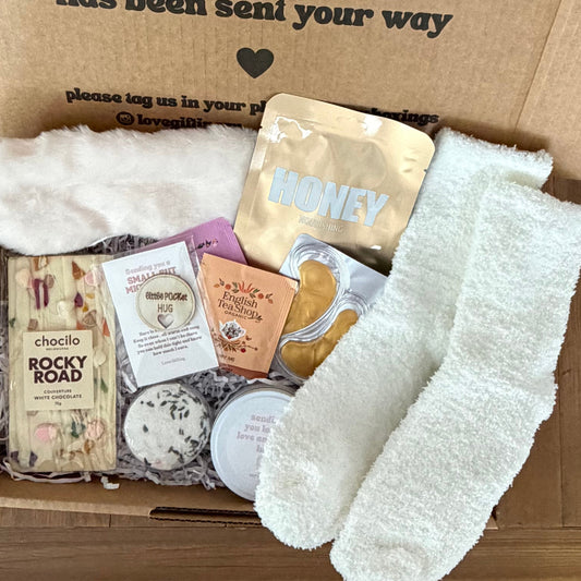 Box of care package with socks, snacks, and a card on a wooden floor.