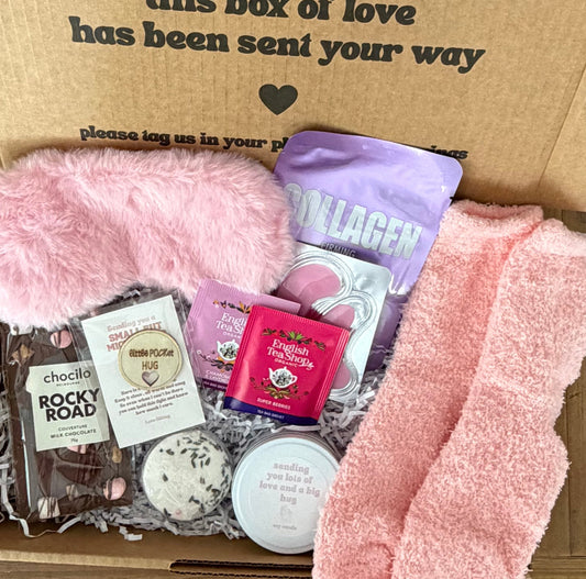 Gift box with pink fuzzy socks, chocolates, and other items on a wooden surface.