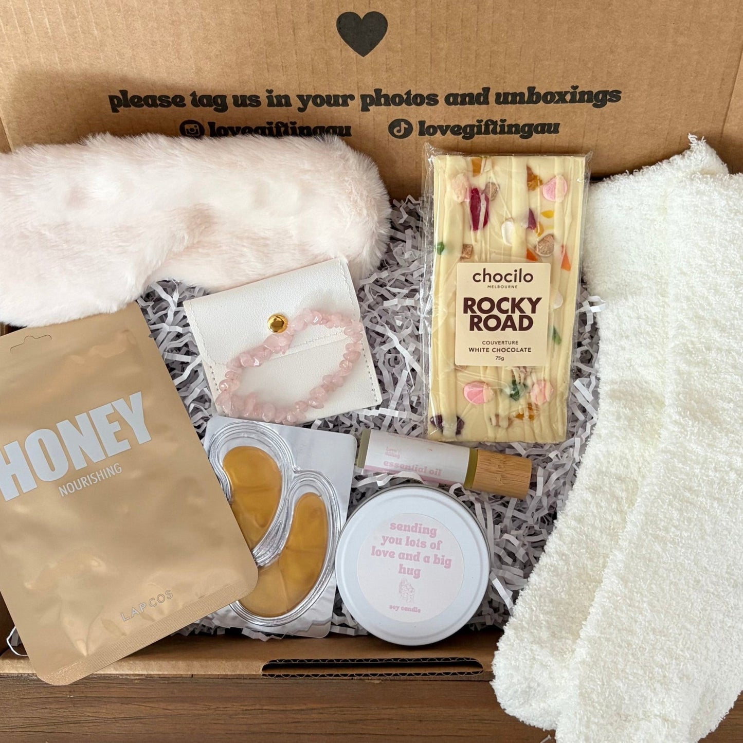 Gifting box with white socks, chocolate bar, and other items on a wooden surface.