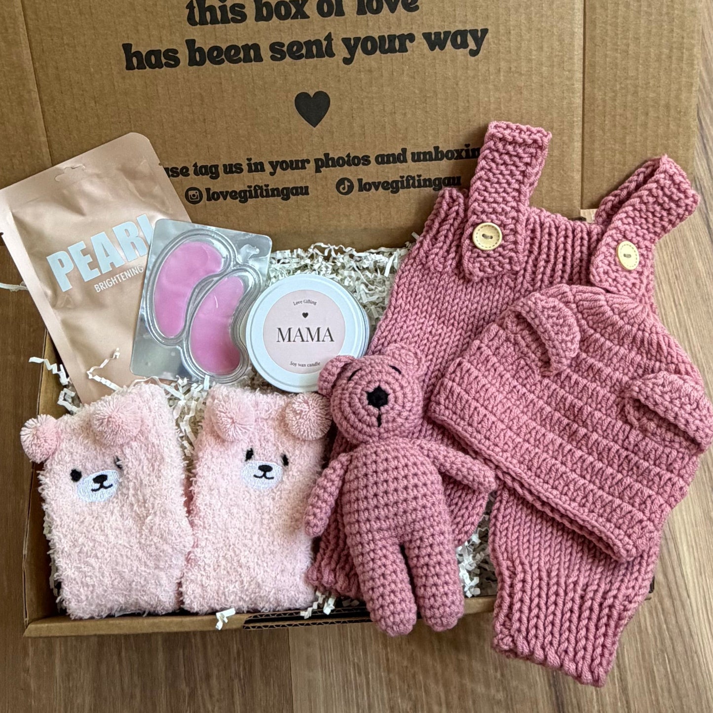 Baby gift set with pink romper, booties, and hat in a box on a wooden floor.