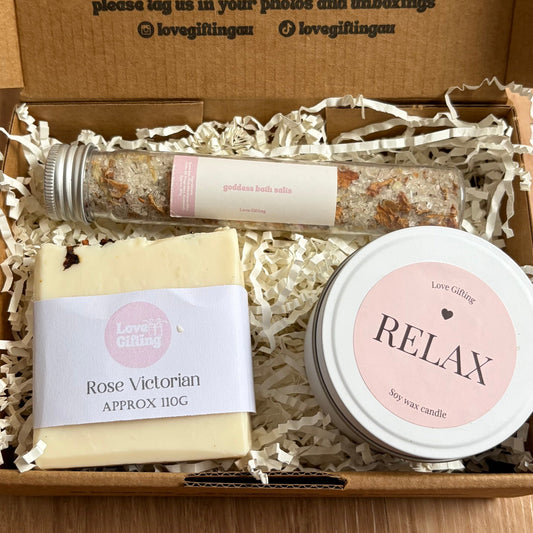Gift box with bath products including a candle, soap, and a small jar labeled 'Relax' on a wooden surface.