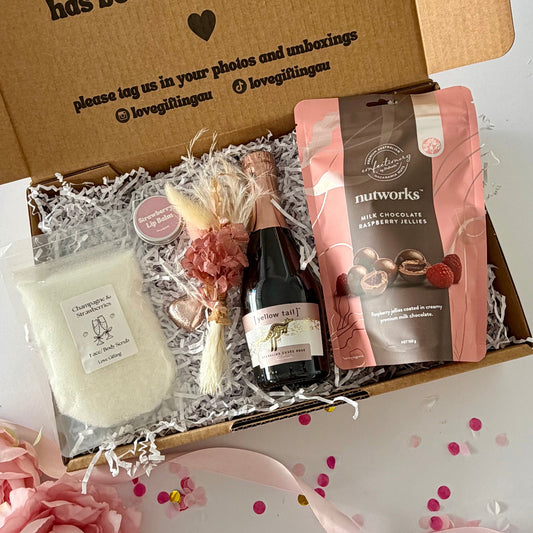 A curated pamper treat gift box s contains a pink pouch of nutworks chocolate-covered raspberries, a small bottle of Yellow Tail Sparkling Rosé, strawberry champagne body scrub, dried flower posy , strawberry lipbalm and a choc heart