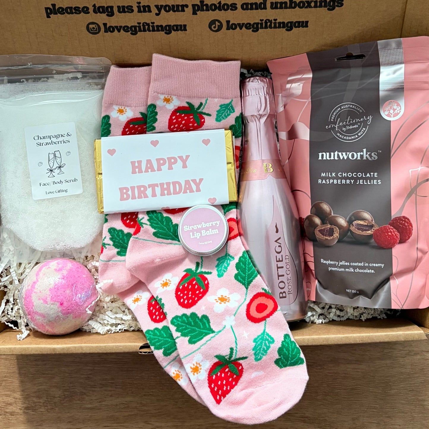 Gift box with birthday-themed items including socks, a bath bomb, and a bag of nuts on a wooden surface.