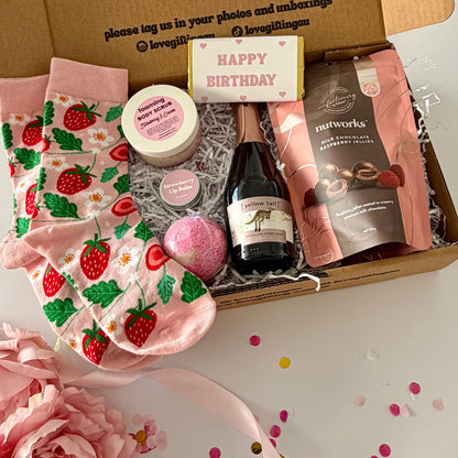 A curated gift box set , pink strawberry socks, a small bottle of cYellow Tail Sparkling Rosé, a jar of foaming body butter, a bath bomb, and a bag of Nutworks chocolates , happy birthday chocolate and strawberry lipbalm
