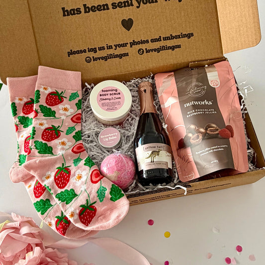 A curated gift box set , pink strawberry socks, a small bottle of cYellow Tail Sparkling Rosé, a jar of foaming body butter, a bath bomb, and a bag of Nutworks chocolates and strawberry lipbalm