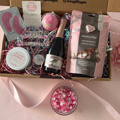 A curated gift box set displayed in an open kraft cardboard box with printed message text inside. The box contains a small champagne bottle, a pink bath bomb, a pink and black bag of snacks, a pink foot file pad, a round pink item, and pink shredded filler.