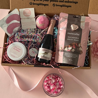 A curated gift box set displayed open on a pink surface, containing a small champagne bottle, a pink bath bomb, a bag of Nutworks chocolate, a pink eye mask, a round bath product, and a jar, with a printed cardboard insert and pink ribbon around it.
