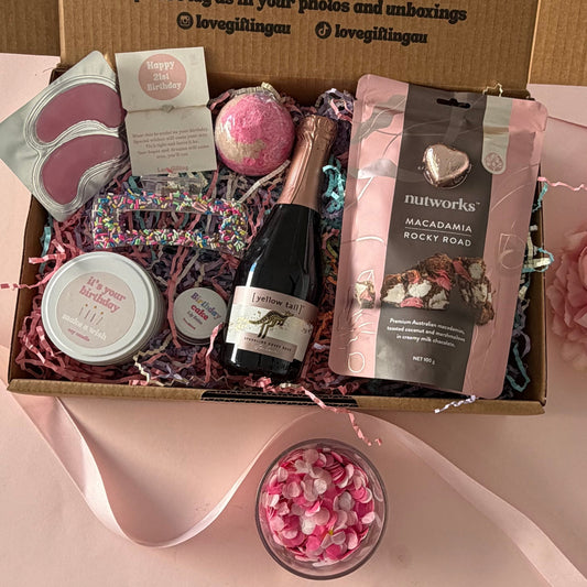 A curated gift box set displayed open on a pink surface, containing a small champagne bottle, a pink bath bomb, a bag of Nutworks chocolate, a pink eye mask, a round bath product, and a jar, with a printed cardboard insert and pink ribbon around it.