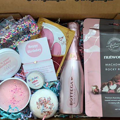 Gift box with various birthday items including a bottle, candles, and a card on a wooden surface.