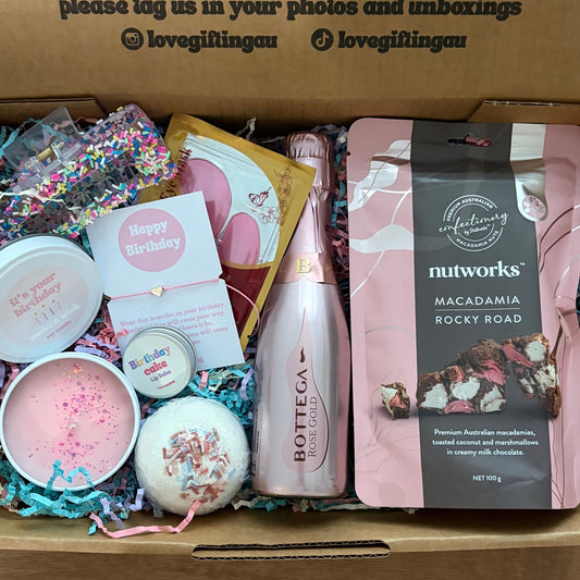 Gift box with various birthday items including a bottle, candles, and a card on a wooden surface.