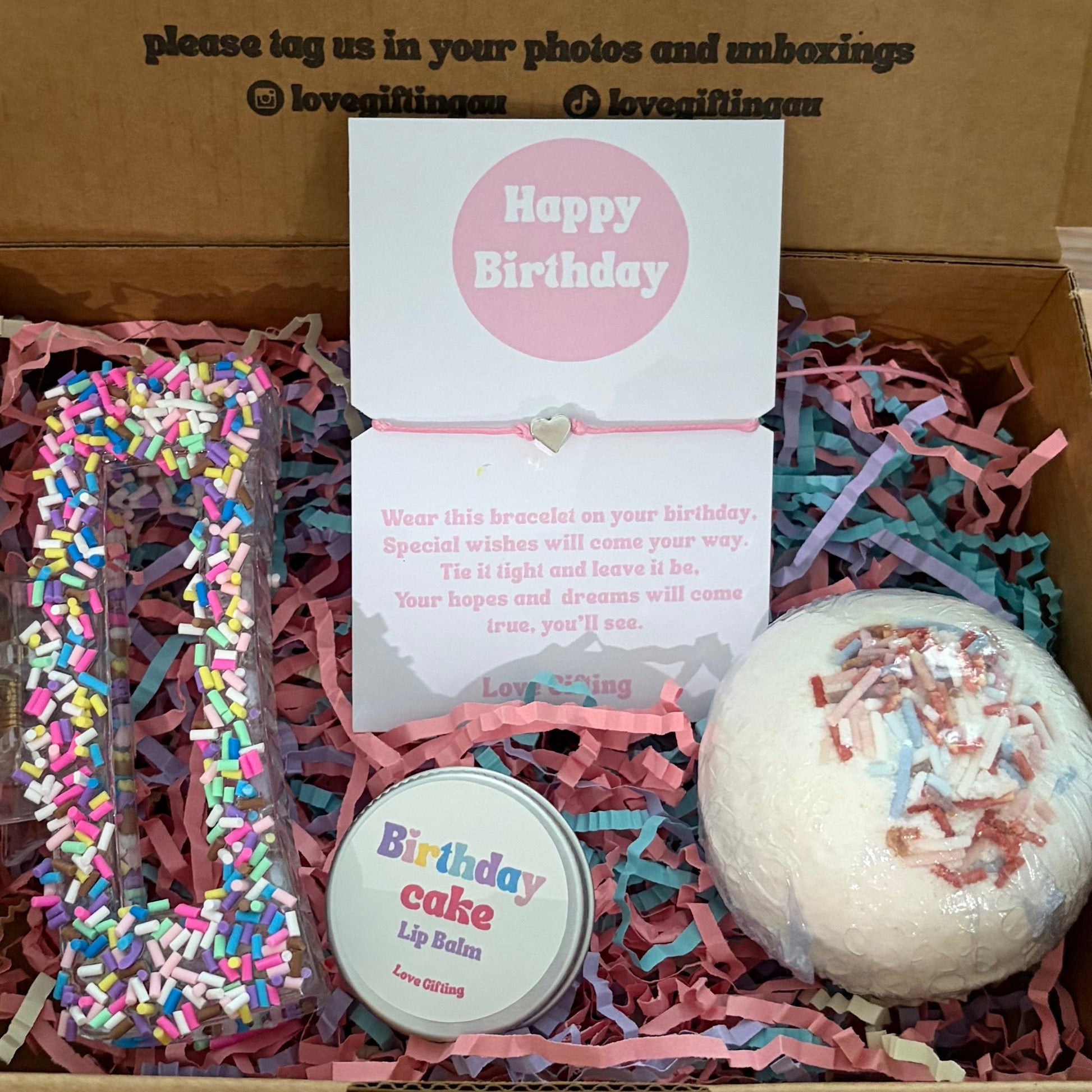 Gift box with birthday-themed items including a bath bomb and cookie-shaped container on a wooden surface.