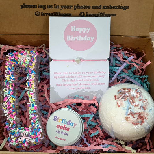 Gift box with birthday-themed items including a bath bomb and cookie-shaped container on a wooden surface.
