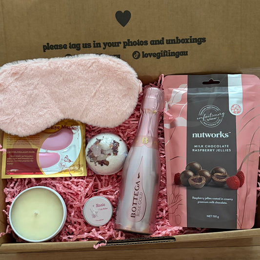 Gift box with various items including a candle, eye mask, and chocolate, on a wooden surface.