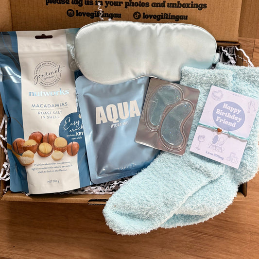 Gift box with items including a sleep mask, towel, and snacks on a wooden surface.