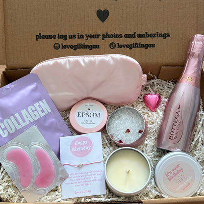 Gift box with self-care items including a bottle, candle, and facial masks on a wooden surface.
