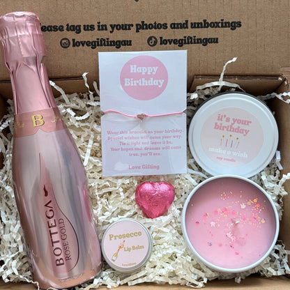 Gift box with pink bottle, card, and candles on a wooden surface
