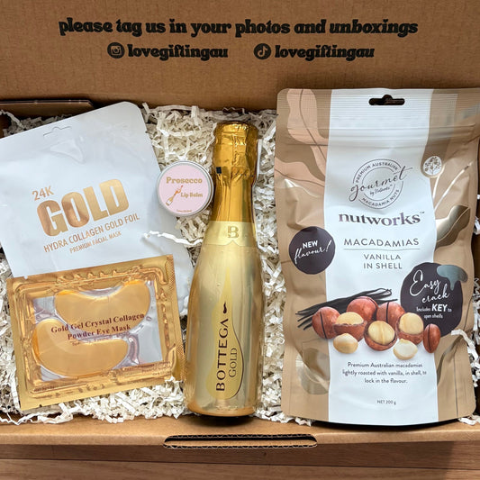 Gift box with a bottle of champagne, snacks, and a card on a wooden surface.