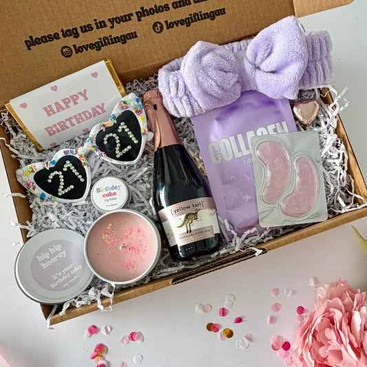 A curated birthday gift box filled with spa and self-care items, photographed from above on a white surface with pink flowers around it. The open kraft box lid has printed text and social handles. Prosecco , happy birthday chocoalte bar, 21st glasses, candle, lapcos facemask, under eye, towelling headband, birthday cake lipbalm, chocolate heart.